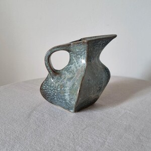 May include: A ceramic jug with a distinctive, angular, and asymmetrical form. The surface displays a mottled, textured glaze in shades of muted blue, green, and grey, with darker brown edges along the rim and handle. It features a wide, curved spout.