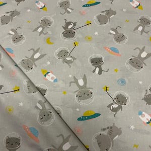 Könnte beinhalten: Grauer Stoff mit einem sich wiederholenden Muster von Cartoon-Katzen in Weltraumhelmen, Raketen, Planeten und Sternen. Die Katzen sind grau mit weißen Bäuchen und halten Zauberstäbe. Die Raketen sind weiß, blau und gelb. Ideal für Kinderprojekte.