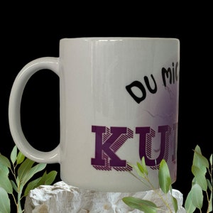 Könnte beinhalten: Weiße Keramiktasse mit einem lila und schwarzen Design. Die Tasse hat den Text "DU MICH" und "KUNI" aufgedruckt.