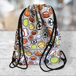 Könnte beinhalten: Eine graue Kordeltasche mit einem Sportball-Druck. Die Tasche zeigt eine Vielzahl von Sportbällen, darunter Basketbälle, Fußbälle, Basebälle und Volleybälle. Die Tasche hat einen schwarzen Kordelzug und einen schwarzen Reißverschluss.