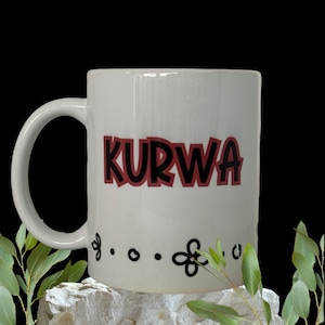 Könnte beinhalten: Eine weiße Keramiktasse mit einem schwarzen und roten Design, auf dem in fetten Buchstaben "KURWA" steht. Die Tasse hat unten einen schwarzen und weißen Blumenschmuck.