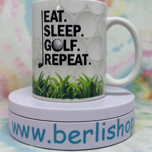 Könnte beinhalten: Eine weiße Keramik-Kaffeetasse mit einer schwarz-weißen Grafik von Golfbällen und dem Text "Eat. Sleep. Golf. Repeat."