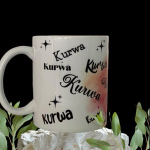 Könnte beinhalten: Eine weiße Keramiktasse mit einem schwarz-weißen Design. Die Tasse hat das Wort "Kurwa" mehrmals in verschiedenen Stilen darauf geschrieben. Die Tasse steht auf einem weißen Stein.
