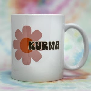 Könnte beinhalten: Weiße Keramik-Tasse mit einem Retro-Blumenmuster in Pfirsich und Orange. Das Wort "KURWA" ist in einer Retro-Schriftart gedruckt. Die Tasse hat einen weißen Henkel. Der Hintergrund ist ein farbenfrohes Batikmuster.