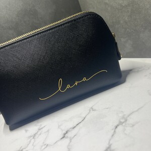 Könnte beinhalten: Schwarze Kosmetiktasche mit goldfarbenem Reißverschluss und dem Namen "Lana" in goldener Schrift.