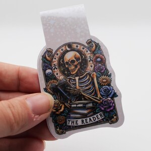 Könnte beinhalten: Ein weißes Lesezeichen mit einer bunten Illustration eines Skeletts, das ein Buch liest. Das Skelett ist von Blumen umgeben und der Text "The Reader" befindet sich unten.