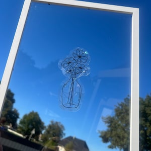 Könnte beinhalten: Ein weiß gerahmter Spiegel mit einer klaren Glasoberfläche. Ein Aufkleber einer Vase mit Blumen ist auf dem Glas angebracht. Der Hintergrund zeigt einen strahlend blauen Himmel und Bäume.