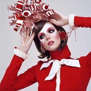 May include: A woman wearing a red sweater with a white bow and white fur trim. She is wearing a festive candy cane themed hat and makeup.