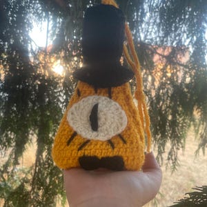 May include: A crocheted yellow triangle with a black top hat and a white eye. The triangle has a black bow tie and is a character from the show Gravity Falls.