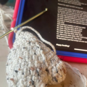 Op de afbeelding: Een close-up van een gehaakt, hartvormig object in wit en zwart gespikkeld. Het object wordt bewerkt met een haaknaald.