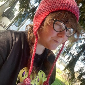 May include: A pink and red crocheted beanie with a pom pom on top and long ties with tassels. The beanie is worn by a person with glasses and a black t-shirt with a yellow graphic.