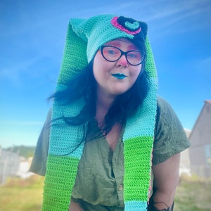 Op de afbeelding: Een persoon draagt een lange, gehaakte sjaal met groene en blauwe strepen en een bijpassende muts met een roze en zwart hartontwerp. De muts heeft een lange, puntige punt.