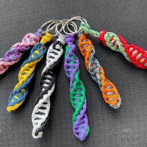 May include: A collection of colorful braided keychains. Each keychain features a unique pattern and color combination, including pink, yellow, black, purple, orange, and red. The keychains are attached to silver metal rings.