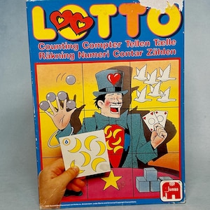 Peut inclure: Boîte de jeu de comptage Lotto vintage avec une illustration de magicien. La boîte présente le mot "LOTTO" en lettres jaunes avec des cœurs rouges. Le jeu comprend des illustrations de balles, de colombes, de cartes et de lunes. La boîte contient également du texte en plusieurs langues.