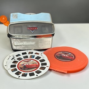 May include: A silver and light blue View-Master toy featuring a Disney Pixar Cars logo on the front, accompanied by three circular white image reels and one orange plastic storage case, each decorated with the red race car character Lightning McQueen.