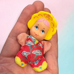 Peut inclure: Une petite poupée avec une coiffe jaune moulée et des cheveux roses. La poupée a des yeux bleus et un visage peint. Elle porte une tenue en tissu rouge avec un motif floral bleu et blanc, agrémentée de sections en tissu jaune sur les pieds.