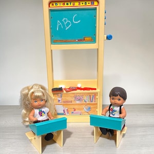 May include: A plastic classroom playset featuring a tall beige chalkboard unit with the letters a b c d e f g and numbers 3+5=8 at the top. Two small dolls sit at individual teal and beige desks, each wearing a white shirt with a colorful graphic.