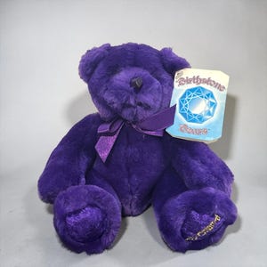 May include: A plush purple teddy bear with soft fur, a dark purple satin ribbon tied around its neck, and a black nose. A tag attached to the ear features the text Birthstone Bears and an illustration of a blue faceted gemstone.