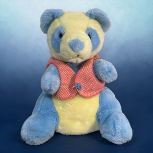 May include: A soft panda bear toy with blue and yellow fur, wearing a pink and orange vest. The panda has blue accents around its eyes, ears, and limbs. The vest is fastened with a blue button.