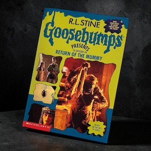 Peut inclure: Un livre Goosebumps Presents intitulé Return of the Mummy par R.L. Stine. La couverture jaune présente du texte bleu, un logo Scholastic et une image centrale d'une momie. Des photos encastrées montrent une personne, un pendentif et un visage.