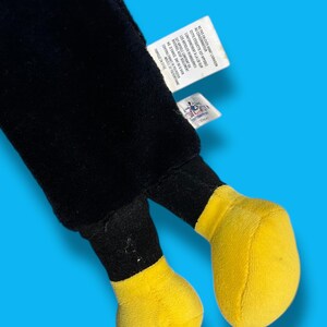 May include: A plush toy featuring black legs and bright yellow feet. The toy has a tag attached with text in French. The background is a solid light blue.