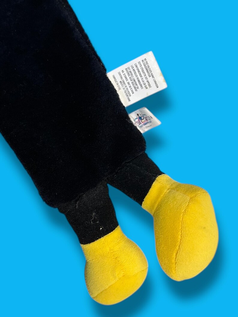 May include: A plush toy featuring black legs and bright yellow feet. The toy has a tag attached with text in French. The background is a solid light blue.