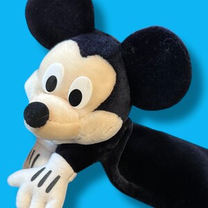 May include: A plush Mickey Mouse toy with black ears, a black body, and white gloves. The face is cream-colored with black eyes and a black nose. The toy is set against a bright blue background.