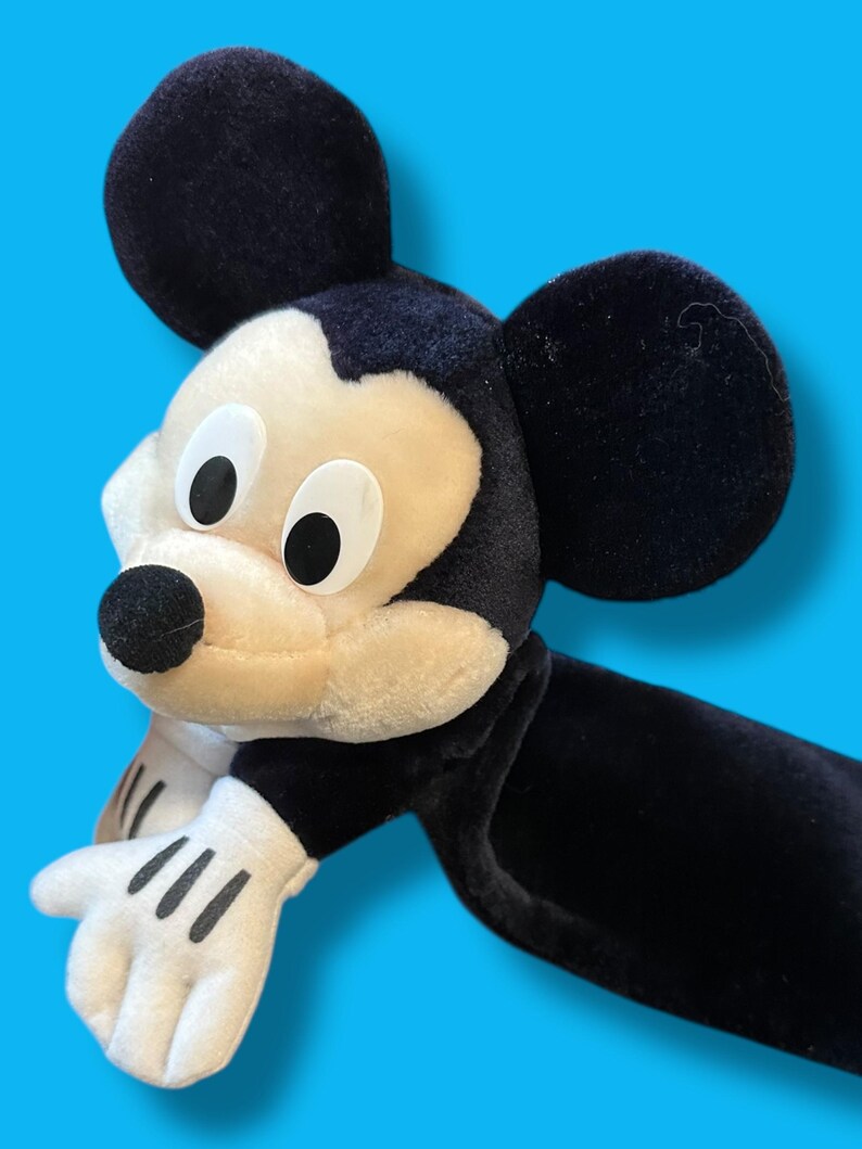 May include: A plush Mickey Mouse toy with black ears, a black body, and white gloves. The face is cream-colored with black eyes and a black nose. The toy is set against a bright blue background.