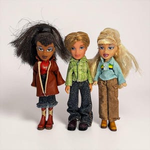 Könnte beinhalten: Drei Bratz-Puppen werden vor einem weißen Hintergrund gezeigt. Die Puppen tragen verschiedene Outfits, darunter eine braune Jacke, einen Jeansrock und Cowboystiefel. Eine weitere Puppe trägt ein grün kariertes Hemd und dunkle Jeans. Die dritte Puppe trägt ein blaues Hemd und eine braune Hose.