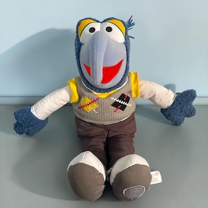 Könnte beinhalten: Ein Plüschtier der Figur Gonzo mit blauem Kopf, langer Nase sowie gelben und weißen Augen. Es trägt eine graue Strickweste mit gelben und schwarzen Flicken, weiße Ärmel, blaue strukturierte Hände sowie eine braune Hose und weiße Schuhe.