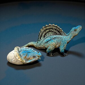 Könnte beinhalten: Zwei Spielzeugdinosaurier werden auf einer dunklen Oberfläche gezeigt. Einer ist ein blauer und beigefarbener Spinosaurus mit einem großen Segel auf dem Rücken. Der andere ist ein Baby-Dinosaurier, das aus einem zerbrochenen Ei schlüpft. Die Spielzeuge sind detailliert und strukturiert.