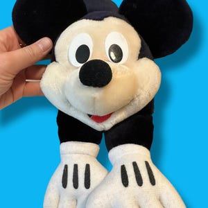 May include: A plush Mickey Mouse toy with large black ears, a black nose, and a red mouth. The toy has white gloves with three black lines on each hand. The background is a solid blue.