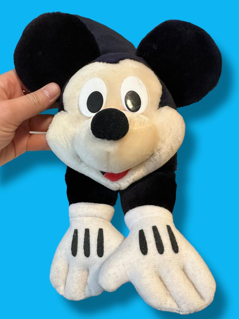 May include: A plush Mickey Mouse toy with large black ears, a black nose, and a red mouth. The toy has white gloves with three black lines on each hand. The background is a solid blue.