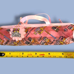 May include: A pink cylindrical pencil case with a repeating cartoon pattern, a top handle, and a flower-shaped charm. A yellow measuring tape placed below shows the case is approximately 8.5 inches long. The text reads Bobby & Kate, Ribbon, and Made in Japan.
