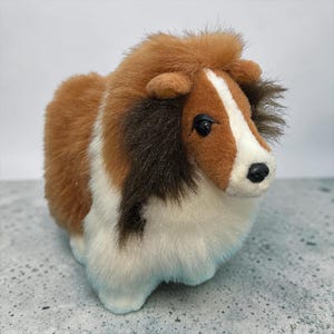 May include: A plush toy dog featuring a tri-colour coat of brown, white, and dark brown fur. The stuffed animal has a white blaze running down its face, dark brown patches around the ears, and a soft, fluffy texture with black plastic eyes and nose.
