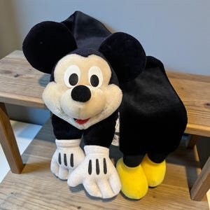 May include: A plush Mickey Mouse scarf with black ears, a white face, and black and white gloves. The scarf has black fabric and yellow shoes. The scarf is resting on a wooden surface.