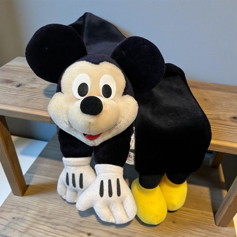 May include: A plush Mickey Mouse scarf with black ears, a white face, and black and white gloves. The scarf has black fabric and yellow shoes. The scarf is resting on a wooden surface.