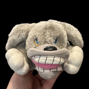 Peut inclure: Un jouet en peluche gris en forme de chien avec un grand sourire édenté et des yeux bleus. Le jouet a des oreilles tombantes et des pattes blanches. La bouche du chien est rose avec des lignes noires. Le fond est noir.