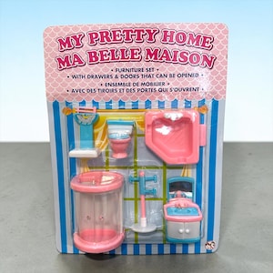 Peut inclure: Un ensemble de mobilier My Pretty Home sous emballage blister comprenant des articles de salle de bain miniatures dont une douche rose, des toilettes, un lavabo avec miroir, un porte-serviettes et une balance. L'emballage porte du texte.