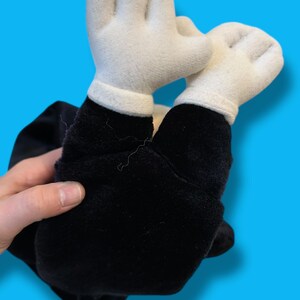May include: A plush toy featuring white gloved hands and black arms. The toy is made of soft fabric and has a classic cartoon aesthetic. The hands are raised, suggesting a playful or celebratory pose.