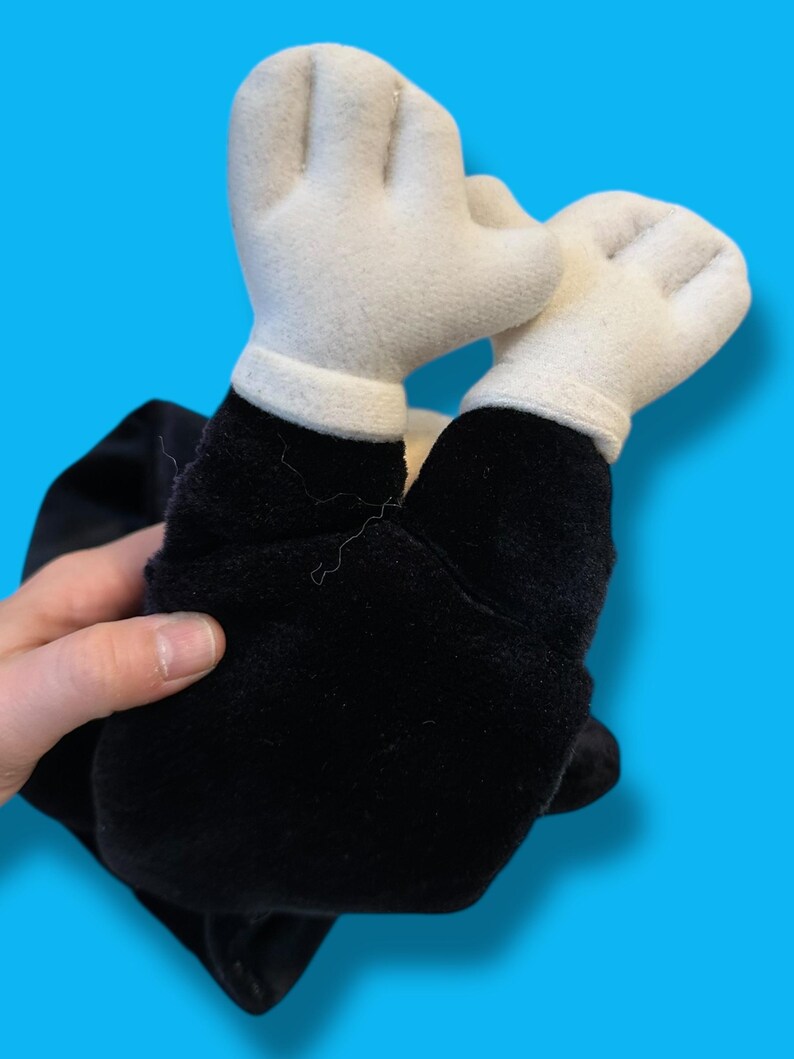 May include: A plush toy featuring white gloved hands and black arms. The toy is made of soft fabric and has a classic cartoon aesthetic. The hands are raised, suggesting a playful or celebratory pose.