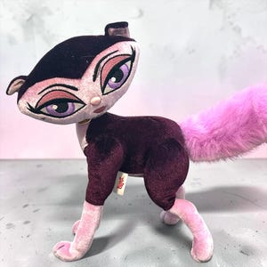 Peut inclure: Une peluche en forme de chat avec un corps en velours violet foncé, des pattes en velours rose clair et une longue queue rose duveteuse. Le chat a un visage stylisé avec de grands yeux violets, du fard à paupières rose et une étiquette Brtaz.