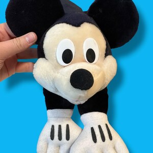 May include: A plush Mickey Mouse toy with large black ears, a black nose, and white gloves with black lines. The toy has a cream-colored face and is set against a blue background.
