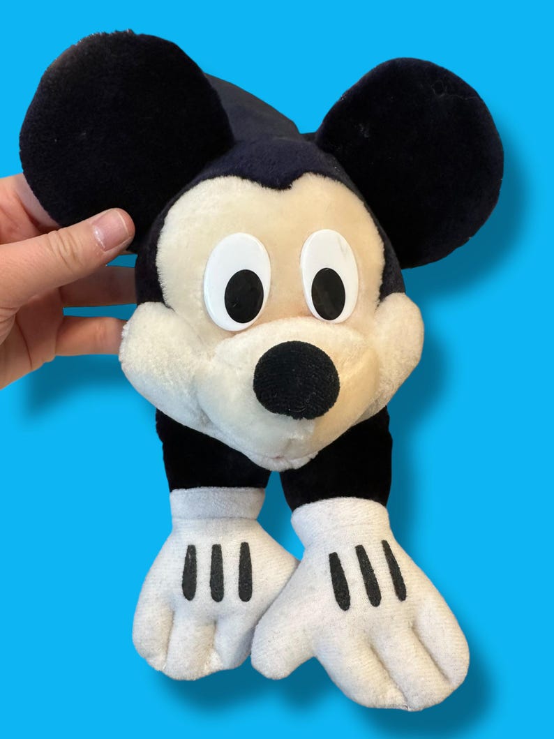 May include: A plush Mickey Mouse toy with large black ears, a black nose, and white gloves with black lines. The toy has a cream-colored face and is set against a blue background.