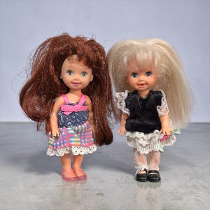 Pode incluir: Duas bonecas pequenas com cabelo comprido. Uma boneca tem cabelo castanho e usa um vestido colorido com um laço rosa. A outra boneca tem cabelo loiro e usa um top preto com uma saia de renda branca. Ambas as bonecas têm traços faciais pintados.