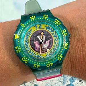 Peut inclure: Une montre-bracelet en plastique bleu sarcelle translucide dotée d'une lunette rotative avec des chiffres jaunes de 5 à 55. Le cadran affiche une couronne de laurier jaune, un grand chiffre 12 bleu et un motif central avec le texte Swatch Swiss.