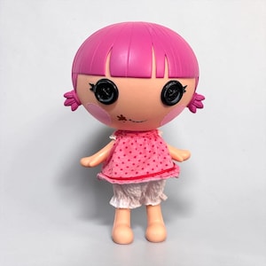 May include: A doll with a pink head and hair, black button eyes, and rosy cheeks. The doll wears a pink polka-dot dress with white trim and white ruffled shorts. The doll's arms and legs are a light peach colour.