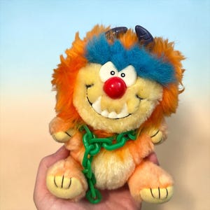 May include: A plush monster toy with orange and yellow fur, a bright blue tuft of hair, and two dark purple horns. The creature has a large red nose, white plastic teeth, and a green plastic chain draped across its chest and front paws.