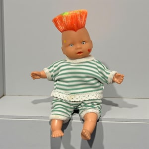 Puede incluir: Muñeca vintage con peinado mohicano naranja y verde. La muñeca viste una camiseta y pantalones cortos a rayas verdes y blancas con ribete blanco. Una estrella y un corazón están pintados en la cara de la muñeca.