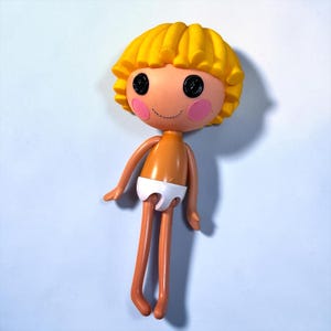 Peut inclure: Une poupée en plastique avec une tête ronde, des cheveux bouclés jaunes et de grands yeux noirs en forme de boutons. La poupée a un visage couleur pêche avec des joues roses et un petit sourire. Elle porte une couche blanche et a de longs bras et jambes fins.